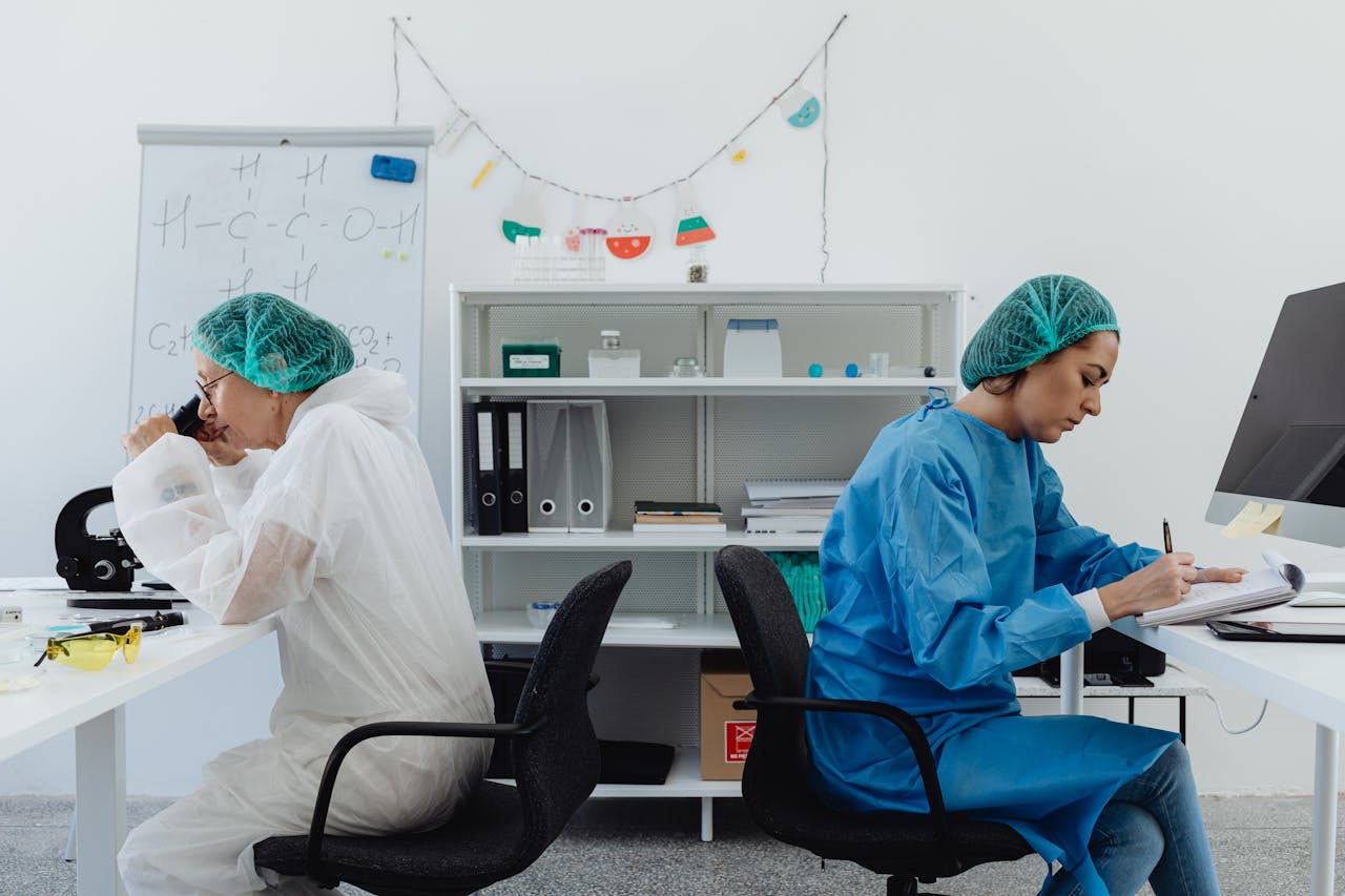 Two scientists in a lab, one examining a microscope, the other taking notes.